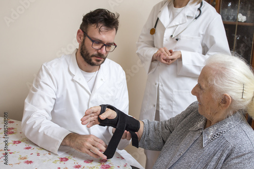 Pielęgniarz w białym kitlu zakłada stabilizator na nadgarstek starej kobiety przy nadzorze lekarza. © Tomasz