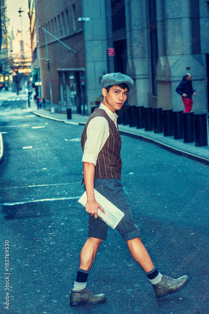 City Boy. Wearing newsboy cap, shirt, patterned vest, gray pants, boot shoes, holding newspaper, Asian American college student walking, crossing narrow street in New York. Filtered Stock-foto | Adobe Stock