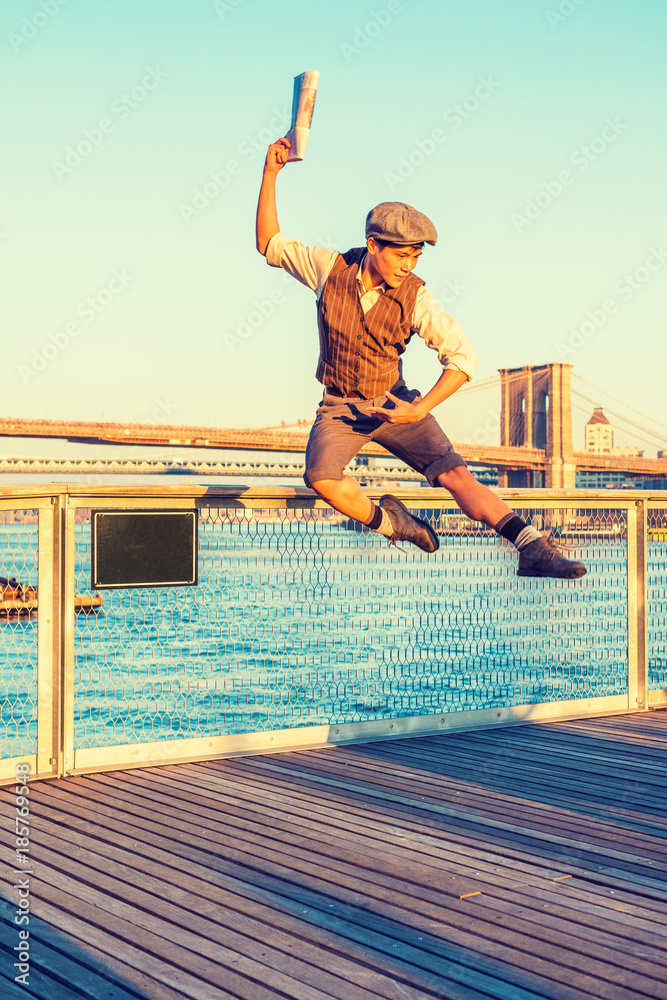 New York City Boy. Wearing newsboy cap, shirt, vest, pants, leather boot shoes, Asian American guy jumping into air at harbor in sunset, raising arm, holding newspaper, dancing with joy, excitement..