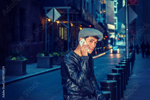 City Life. Wearing black leather jacket, newsboy cap, Asian American college student sitting on narrow street in New York, talking on his cell phone, a car on background. Filtered effect..