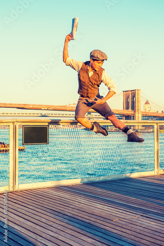 New York City Boy. Wearing newsboy cap, shirt, vest, pants, leather boot shoes, Asian American guy jumping into air at harbor in sunset, raising arm, holding newspaper, dancing with joy, excitement.. photo