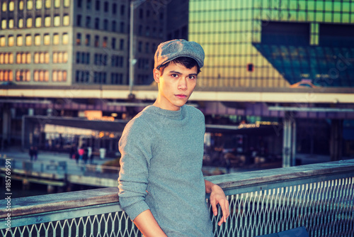 Portrait of New York City Boy. Wearing newsboy cap, knitting sweater, Asian American college student standing in business district with high buildings, looking at you. Filtered effect..