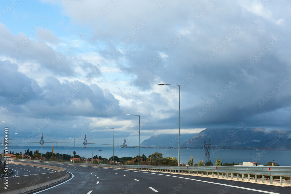 Road along the sea against a blue sky with clouds and a suspension Rion-Antirion Bridge, Gefira Charilaos Trikoupis, Greece