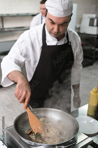 Concentrated chef works at restaurant kitchen. Focused male cook is preparing a meal in the frying pan. Successful professional lifestyle