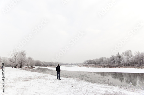 Man in a black coat standing on a white snowy shore of a river. Winter day with frozen weather. Human and strong nature.