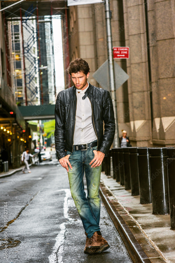 European businessman traveling in New York. Wearing black leather jacket,  white undershirt, blue jeans, brown boot shoes, a young guy with beard  walking on narrow wet street after raining, . Stock Photo