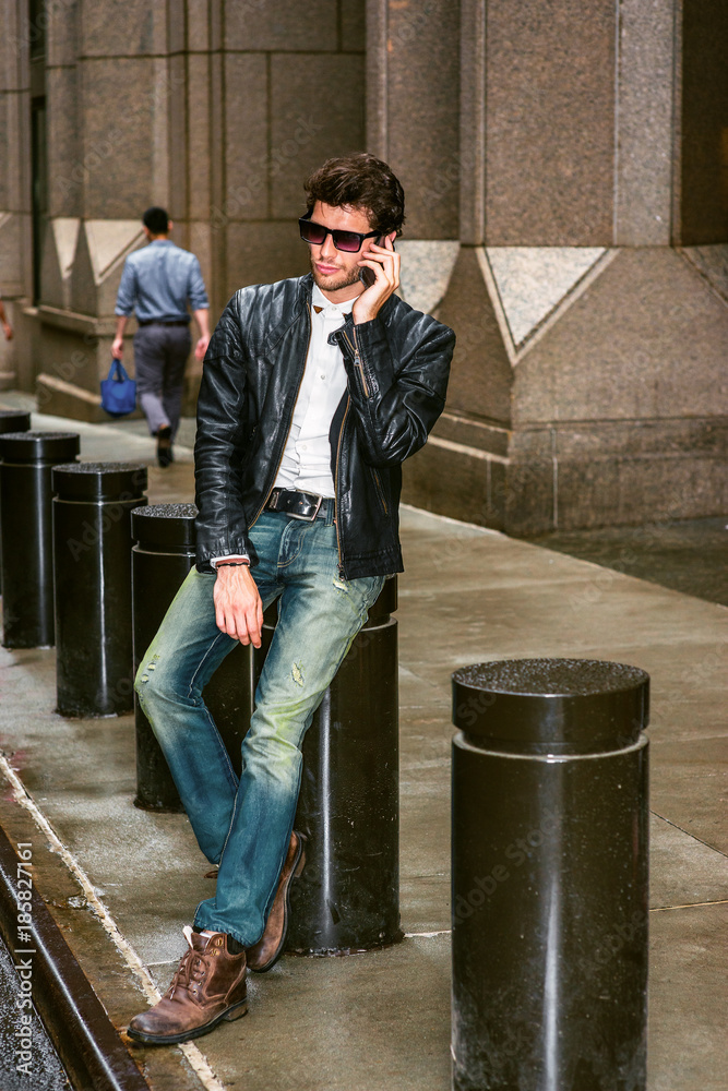 Dressing in black leather jacket, blue jeans, brown boot shoes, wearing  sunglasses, a young European Businessman with beard sitting on street in  New York, talking on mobile phone during working break Stock