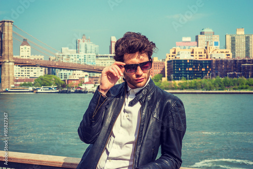 European businessman traveling in New York. Wearing leather jacket, holding sunglasses, a guy with beard, standing by river, looking down, sad, think. Brooklyn bridge, high buildings on background..