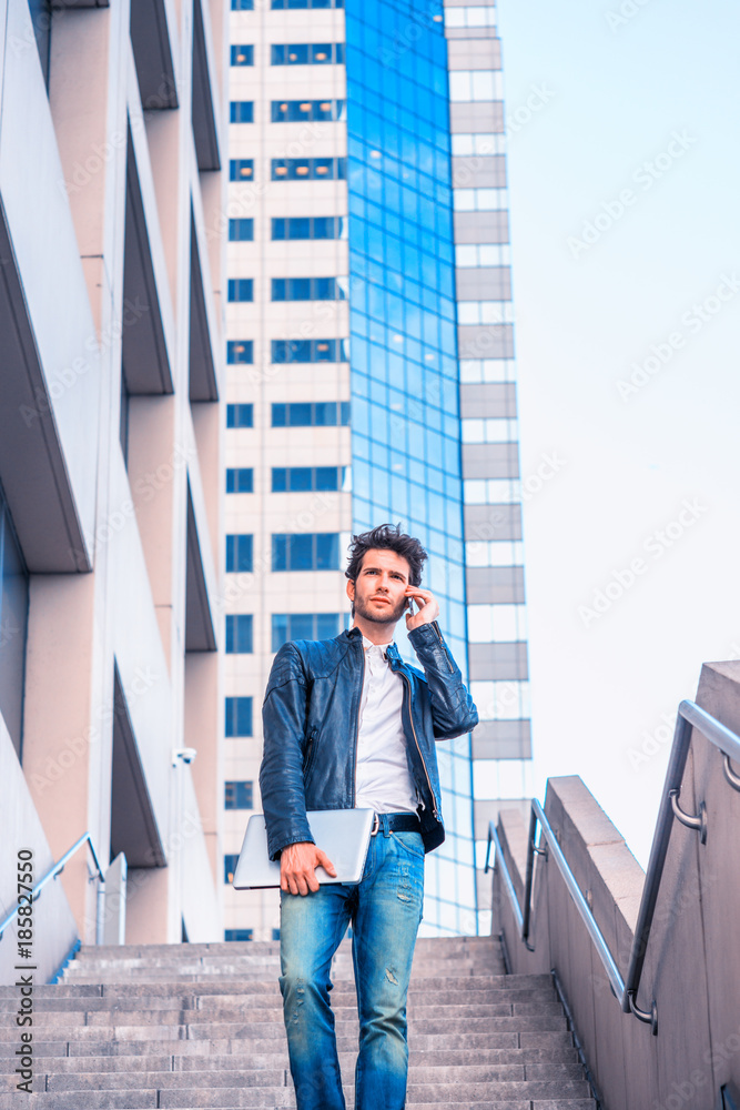 Young professional, wearing leather jacket, jeans, arm carrying laptop computer, talking to his family on his mobile phone, walking down stairs outside office building after working, going home. .