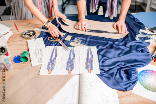 Cutting blue fabric on the table full of tailoring tools. Close-up view on the hands, fabric and fashion drawings photo