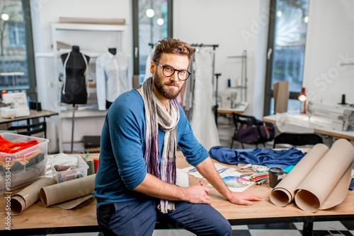 Portrait of a handsome fashion designer sitting with paper sketches at the studio full of tailoring tools and clothes