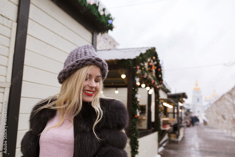 Street portrait of cheerful blonde woman wearing knitted hat and fur coat. Empty space
