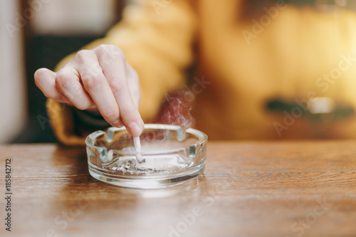 Focus on caucasian young woman hand putting out cigarette on glass ashtray on wooden table, cigarette butt, smoking is dying. Quit smoking. Health concept. Close up photo. photo