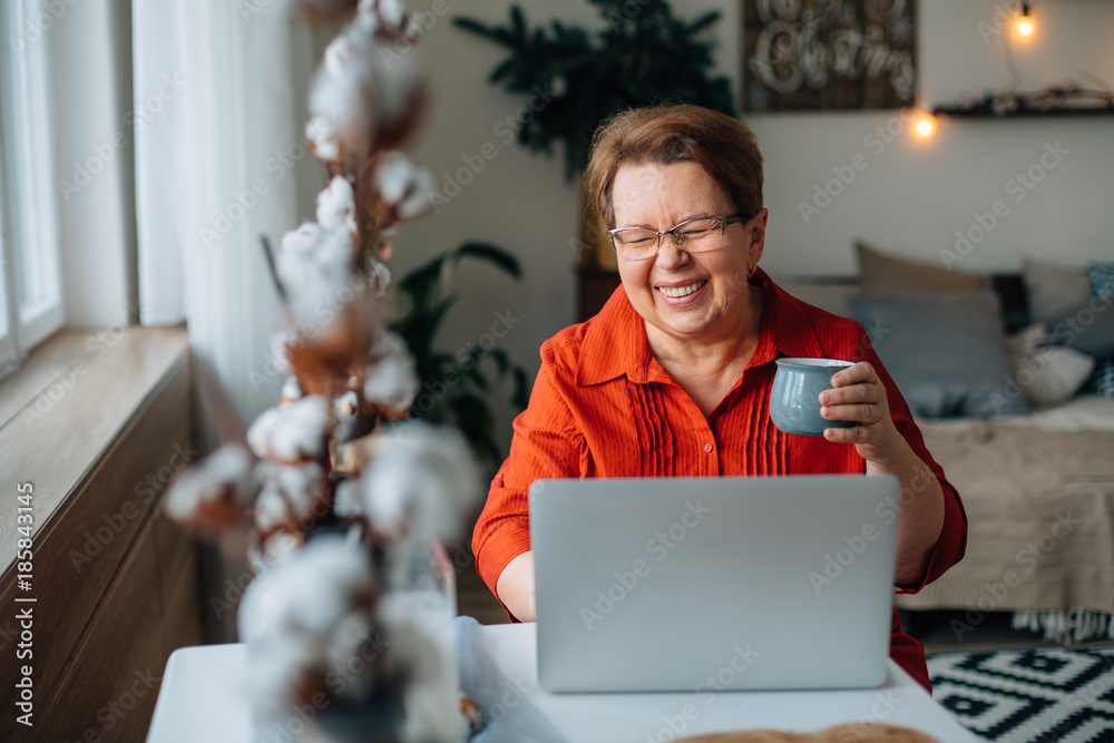 laughing Senior woman using laptop for websurfing. Mature lady with cup of coffee watching funny movie.