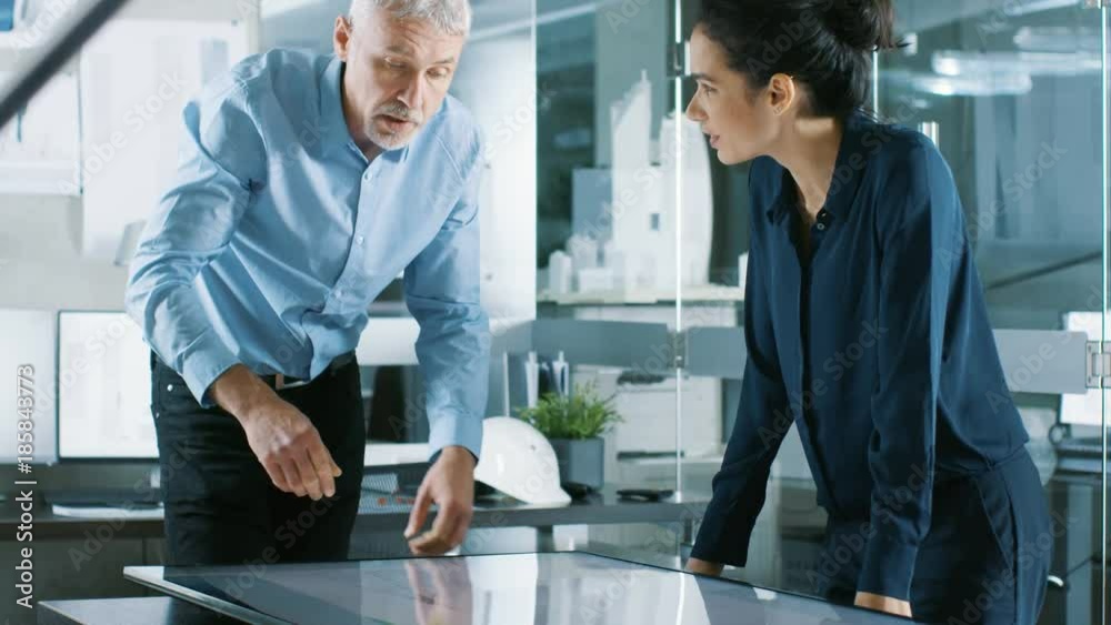Senior Male and Young Female Architectural Designers Draw Building Concept on a GraphicsTablet Display Vertical Touchscreen Table. They Use Gestures for Zooming Project Model. Senior Male and Young Female Architectural Designers Draw Building Concept on a GraphicsTablet Display Vertical Touchscreen Table. They Use Gestures for Zooming Project Model.