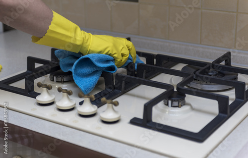 Women's hand in protective gloves wipes the kitchen gas stove with a blue rag.