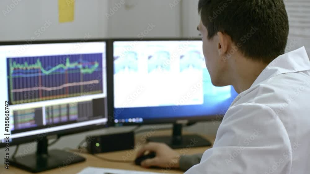 PAN of busy young male scientist in lab coat looking at computer screens with line charts and monitoring activity during experiment