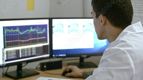 PAN of busy young male scientist in lab coat looking at computer screens with line charts and monitoring activity during experiment