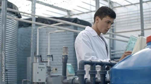 PAN of young male agrochemist in lab coat operating fertigation machine in industrial greenhouse with hydroponic system photo