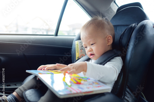 Cute little Asian 18 months / 1 year old toddler baby boy child sitting in car seat holding and enjoy reading book, Happy traveling with child concept, Little Traveler safety, road trip concept