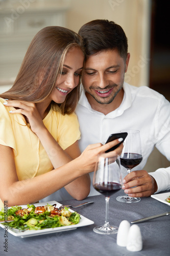 Couple In Love Looking On Phone Having Dinner In Restaurant.