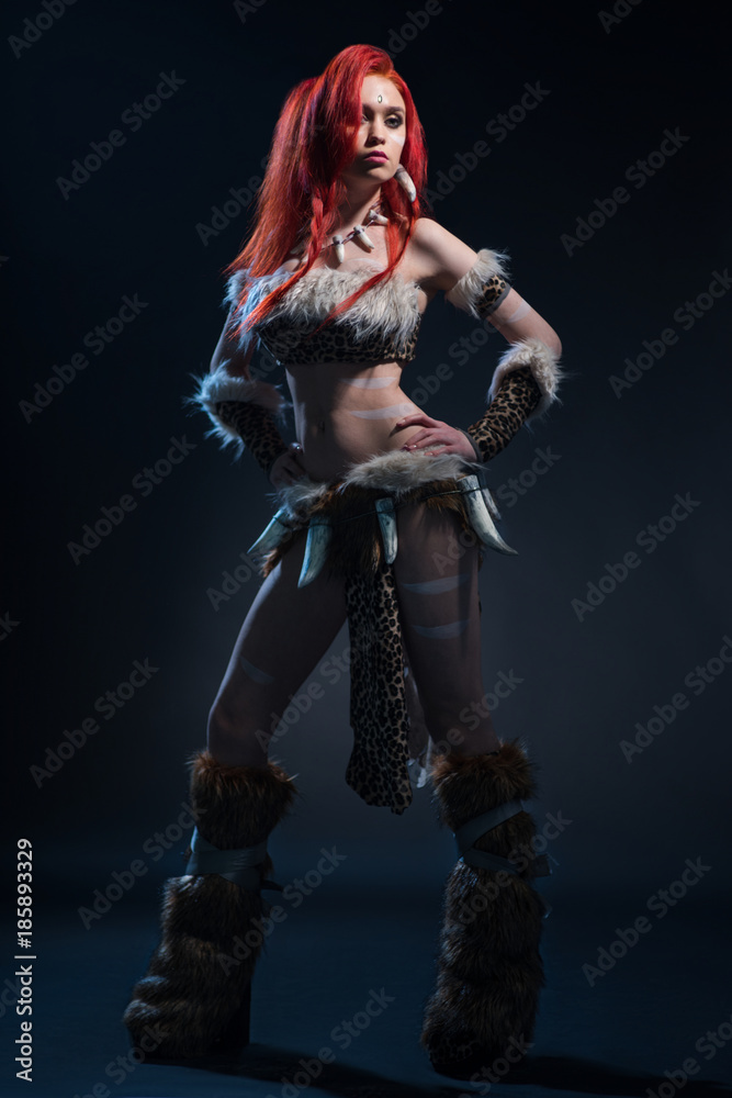 Beautiful red haired woman in stone age clothing with spear posing at camera on dark background - obrazy, fototapety, plakaty 