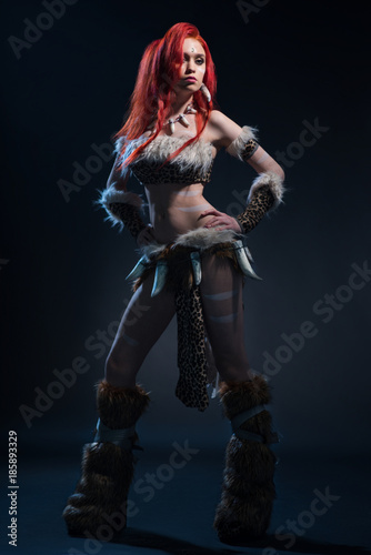 Bild auf Leinwand Beautiful red haired woman in stone age clothing with spear posing at camera on