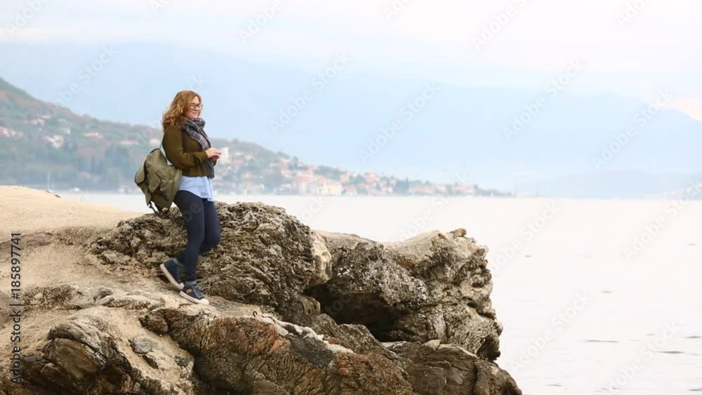 A woman traveler with a backpack enters the frame, stops on a rock and throws coins into the sea. A woman traveler with a backpack enters the frame, stops on a rock and throws coins into the sea.