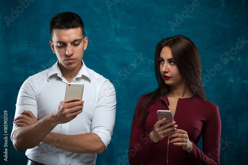 Communication and jealousy. Young beautiful woman with smartphone jealously looks at her husband typing text on smartphone on blue background photo