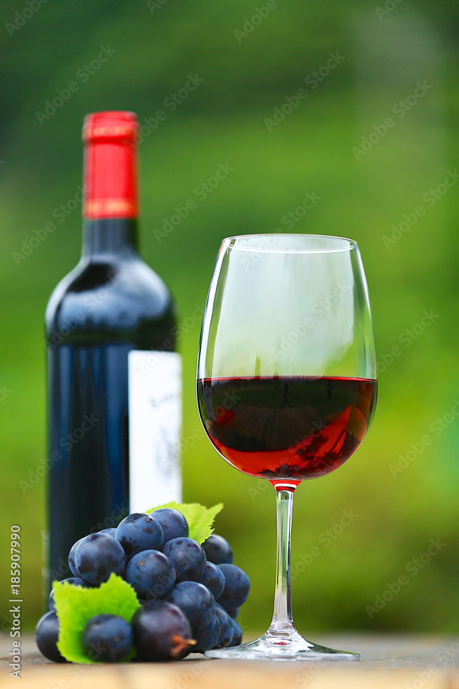 Single Red Wine in Bordeaux-shaped glass with bottle and fresh grapes on wooden table, defocused green outdoors background 1