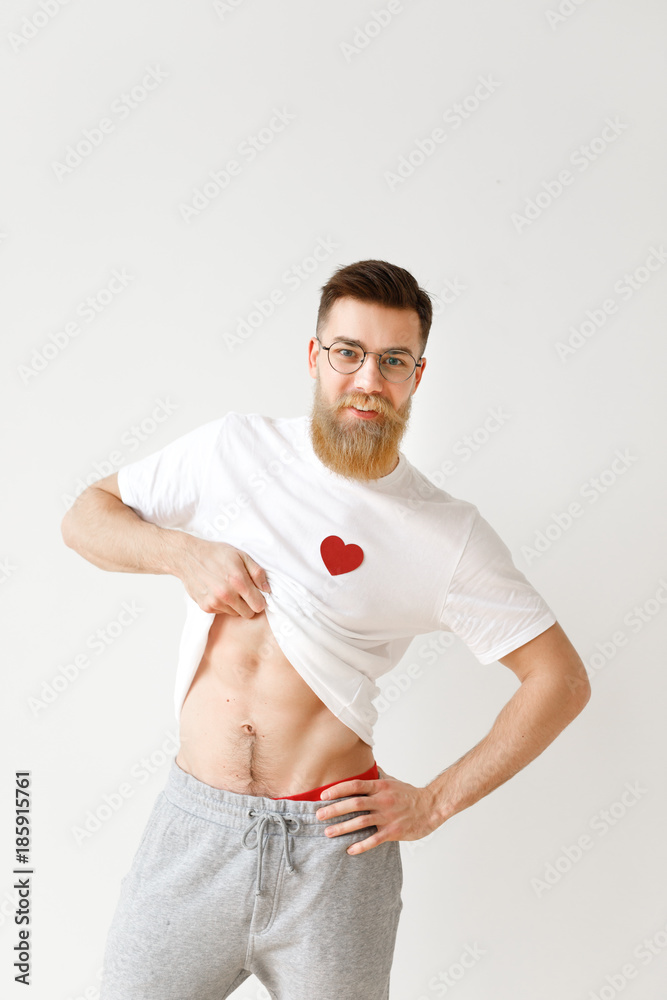 Handsome muscular man raises his white t shirt, demonstrates naked body, poses against white background. Positive bearded male in spectacles shows perfect masculine body. Vertical shot Stock Photo | Adobe