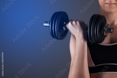 A sporty girl holds a dumbbell in her hands, shakes a muscular. Against a dark background
