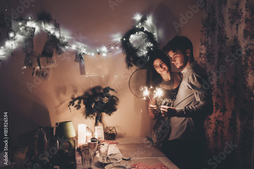 happy new year and merry christmas concept. happy stylish hipster couple holding burning sparklers fireworks and embracing in room at hoome in evening. happy holidays. toned photo