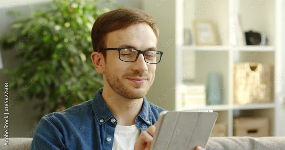 Portrait shot of the attractive young man in glasses and jeans shirt scrolling on the white tablet device in the cozy white room. Indoor Portrait shot of the attractive young man in glasses and jeans shirt scrolling on the white tablet device in the cozy white room. Indoor