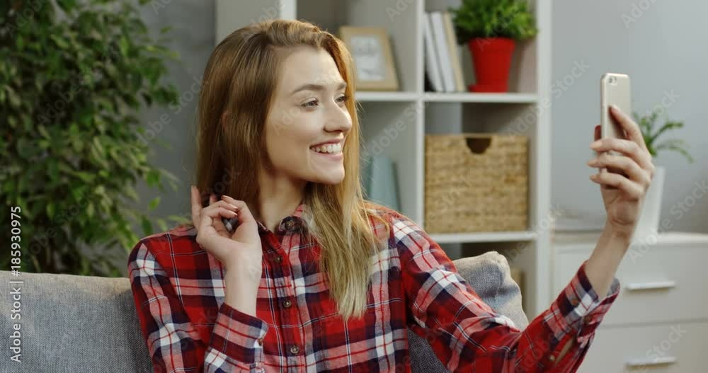 Attractive young blonde woman in the motley shirt making selfies and showing victory gesture while sitting on the couch in the nice room. Portrait shot. Indoors