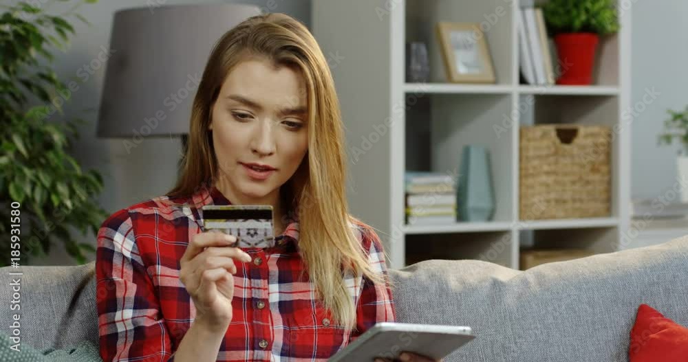 Portrait of the young attractive woman in the plaid shirt buying online and entering credit card data on the tablet device while sitting on the sofa in the cozy room. Inside