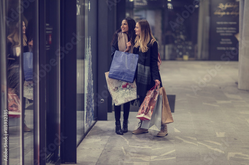 Two cute young adolescent women, heavy loaded with christmas gifts,  shopping, while walking along store windows and choosing and admiring the items. 