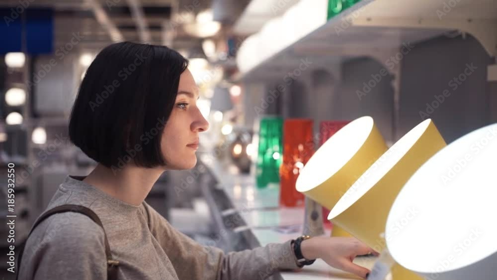 Customer chooses bedroom goods in hypermarket. Young woman examining lamp