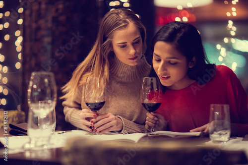 Two adolescent women going through the menue  in a fancy restaurant  while drinking a glass of red wine and laughing and talking  maybe gossiping