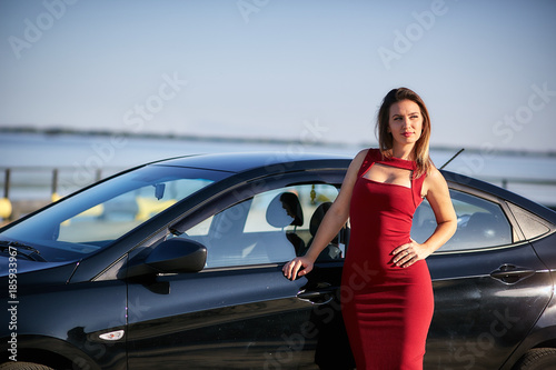 Summer portrait of stylish blonde vintage woman with long legs posing near red retro car. Fashionable attractive fair hair female with black hat near a red vehicle. Sunny bright colors, outdoors shot. © vadiar