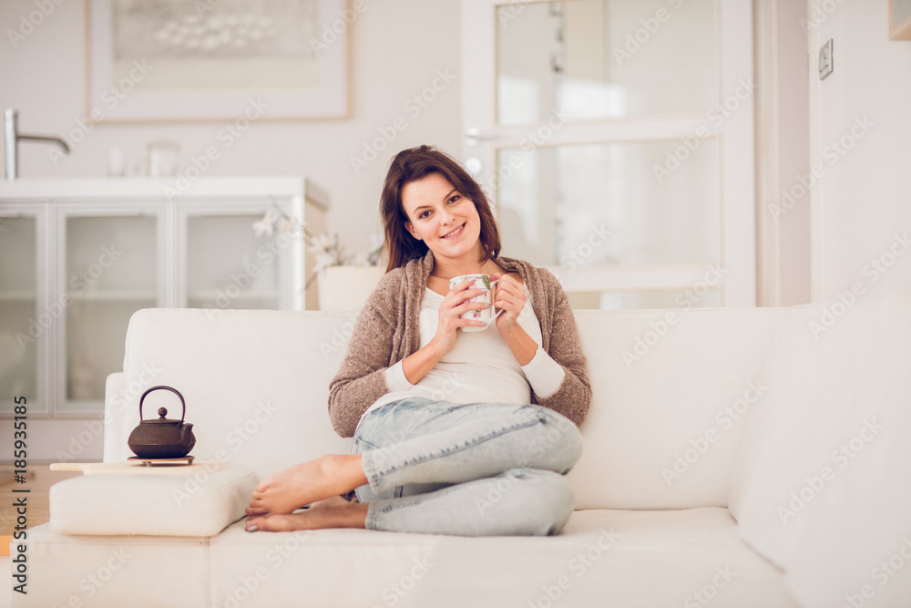 young woman in her late twenties sitting and laying on a lether white sofa in a cosy interier of her bright home and drinking a cup of tea and reading a book, totaly relaxed young woman in her late twenties sitting and laying on a lether white sofa in a cosy interier of her bright home and drinking a cup of tea and reading a book, totaly relaxed
