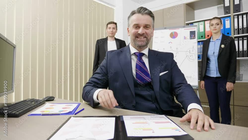 Portrait of businessman working in his busy office with his two colleagues in the background. Senior business person in sitting at the table Portrait of businessman working in his busy office with his two colleagues in the background. Senior business person in sitting at the table
