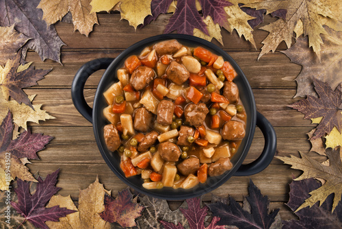 Overhead photo of a pot of beef stew on a wooden table and autumn fall leaves in the background.