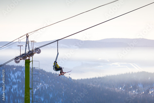Two men skiers in warm clothes and with mountain skis climb up the ski lift up the mountain in a ski resort on a warm winter day, side view