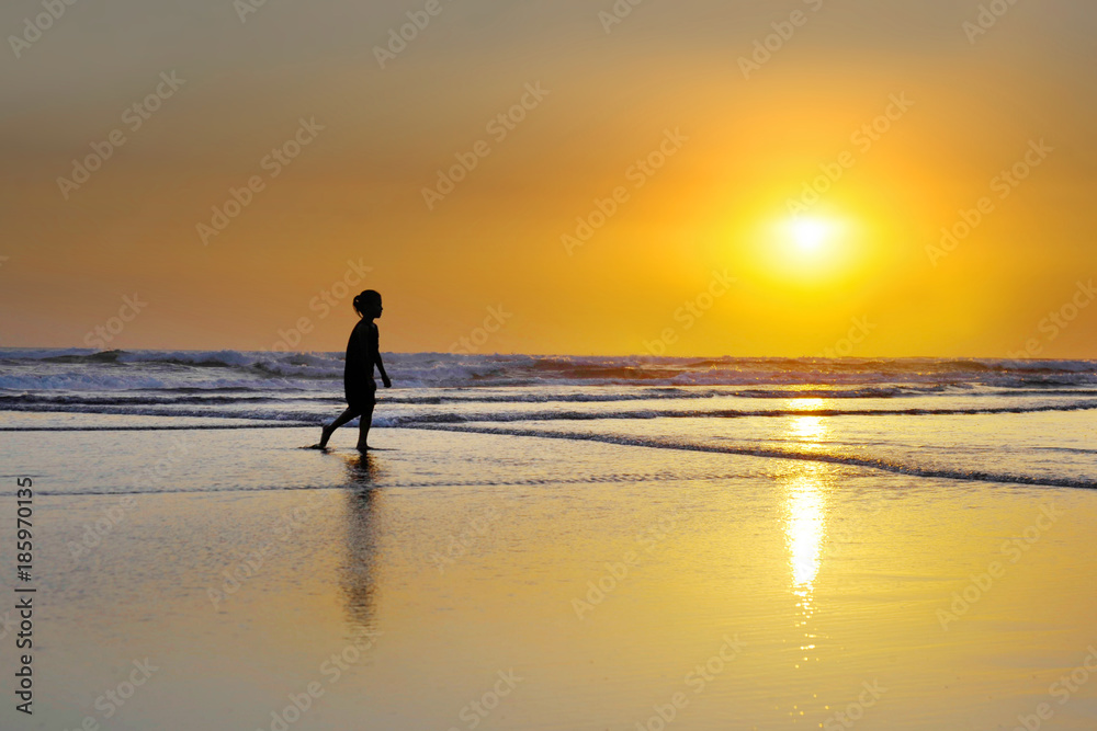 silhouette of anonymous unknown young girl walking solitary on the beach on wet sand with amazing beautiful sunset under orange sky
