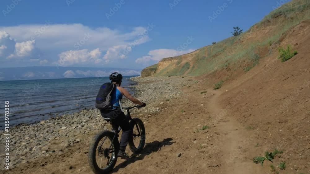 Fat bike also called fatbike or fat-tire bike in summer driving on the road. The guy rides by the hill on a sand clay path, behind him the shore by the sea. Slow motion shooting 180fps. Fat bike also called fatbike or fat-tire bike in summer driving on the road. The guy rides by the hill on a sand clay path, behind him the shore by the sea. Slow motion shooting 180fps.