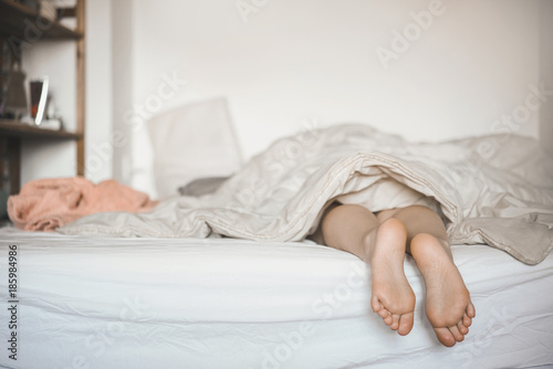 Female legs from under a white blanket on a white bed. The girl is sleeping early in the morning.