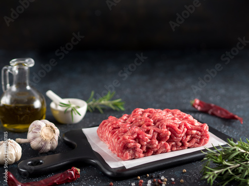 Fresh raw minced beef on backing paper and cutting board and ingredients over black cement background with copy space.