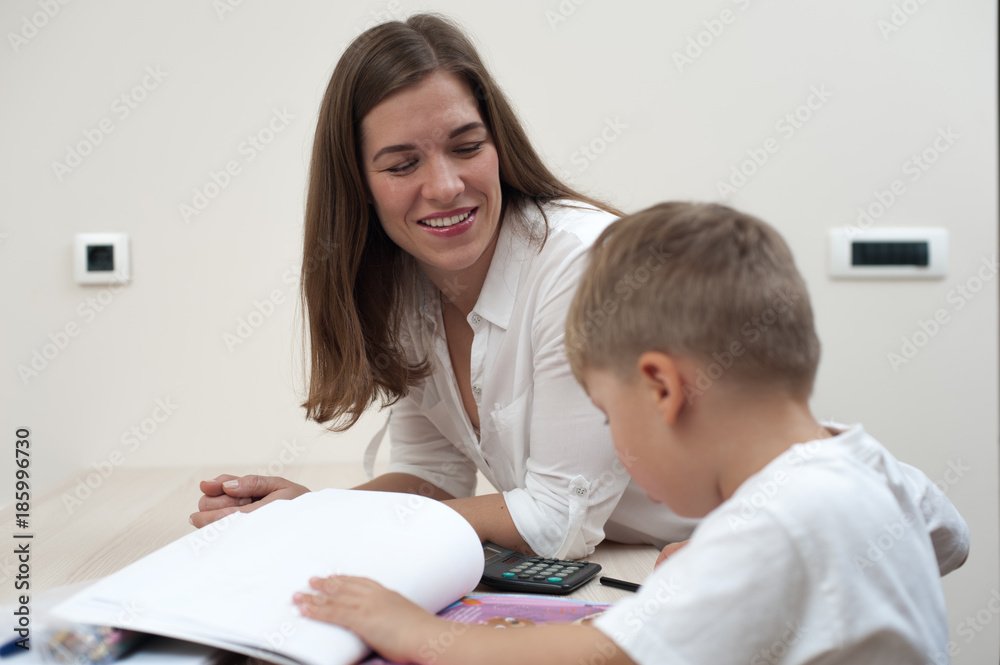 Positive young mother engaged at home with her child sitting at table with books Positive young mother engaged at home with her child sitting at table with books