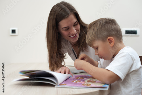 Happy young mother is studying with her young son sitting at the table at home